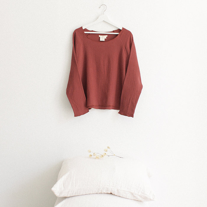 Red brown muslin shirt with long sleeves hanging above a stack of pillows.