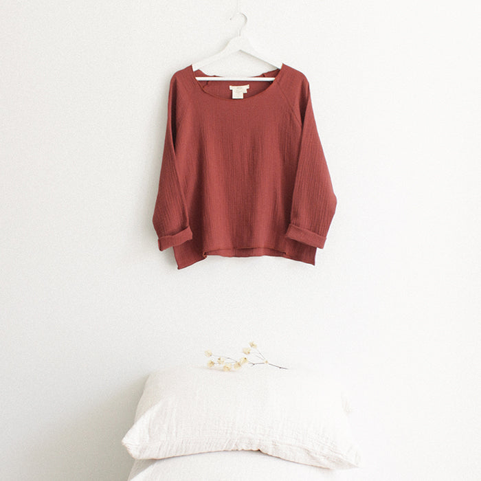 Red brown muslin shirt with cuffed long sleeves hanging above a stack of pillows.