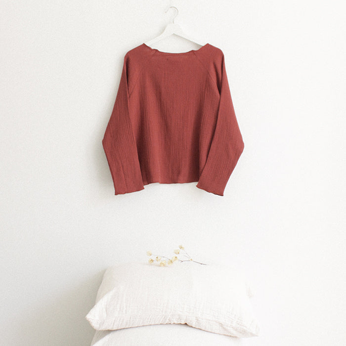 Red brown muslin shirt with long sleeves from the back hanging above a stack of pillows.