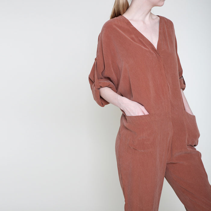 Close up of a woman wearing a rust red long sleeved jumpsuit with her hands in the patch pockets.