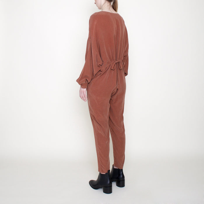 Woman wearing a rust red long sleeved jumpsuit with long legs and buttons from the back side.