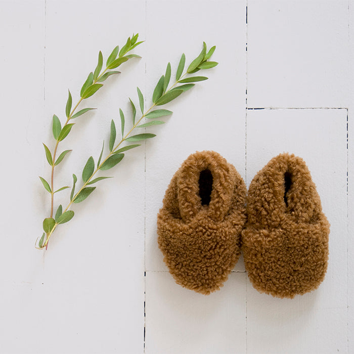 Pair of brown faux shearling baby booties next to some leaves.