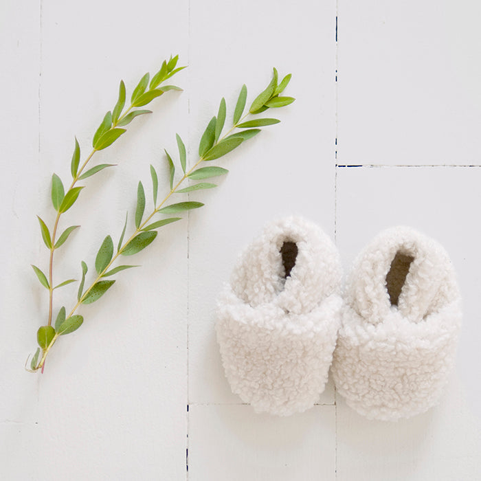 Pair of cream faux shearling baby booties next to some leaves.