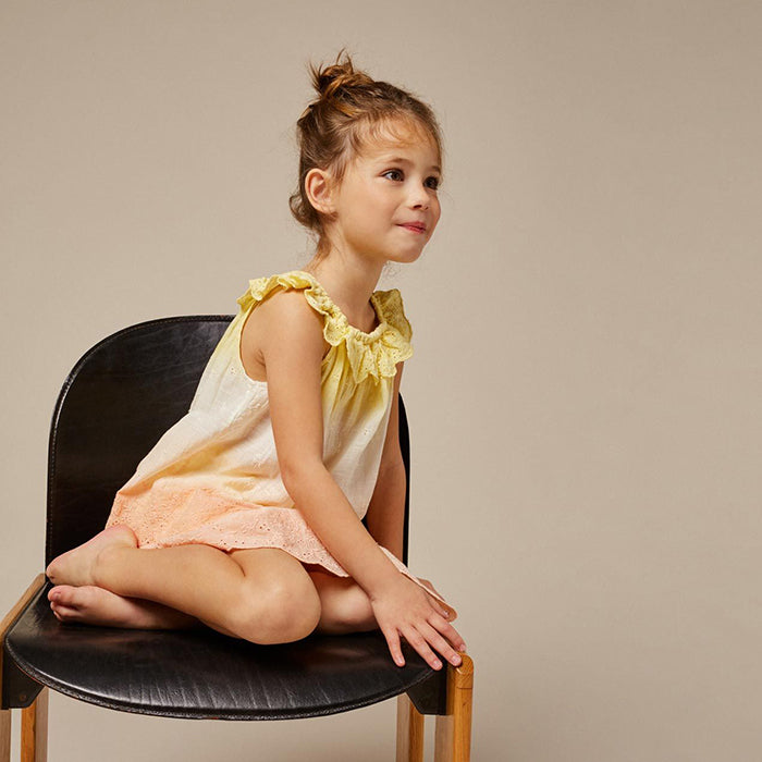 Girl sitting in a chair wearing a sleeveless cotton broderie anglaise blouse in a yellow to pink gradient.