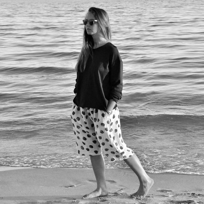 Woman standing on a beach wearing a black long sleeved sweater and polka dot long shorts.