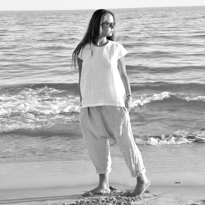 Woman standing on a beach wearing a white blouse and light grey harem pants.