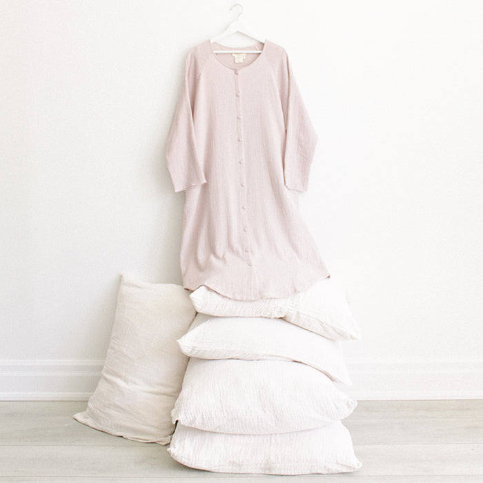 Light pink muslin dress with buttons down the front and long sleeves hanging above a stack of pillows.