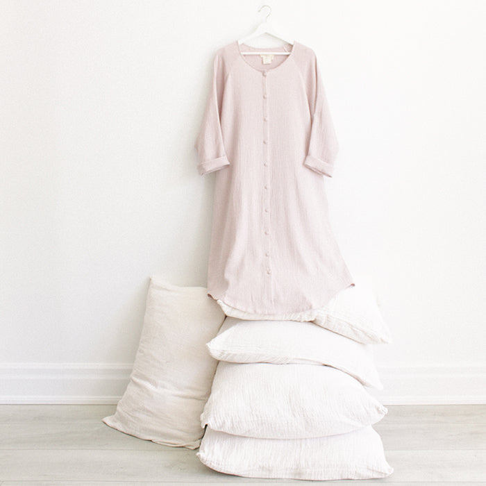 Light pink muslin dress with buttons down the front and cuffed long sleeves hanging above a stack of pillows.