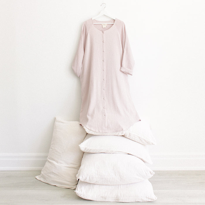 Light pink muslin dress with buttons down the front and long sleeves with a hand in the pocket hanging above a stack of pillows.