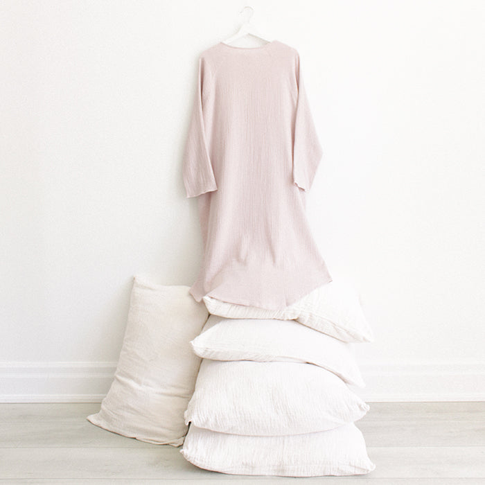 Light pink muslin dress with buttons down the front and long sleeves from the back hanging above a stack of pillows.