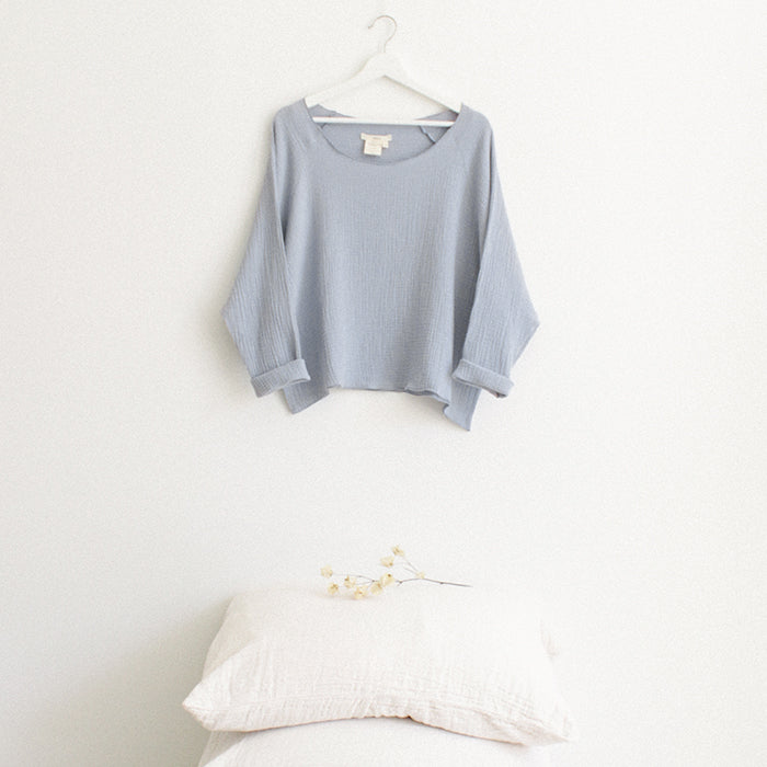 Light blue muslin shirt with cuffed long sleeves hanging above a stack of pillows.